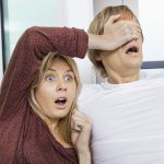 Shocked woman covering man’s eyes while watching TV at home